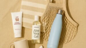 Summer Essentials Kit arranged on sunlit sand, featuring SPF 50 mineral sunscreen, a bamboo beach towel, insulated light-blue water bottle, organic aloe vera gel, mesh tote bag, beige coffee mug, and SPF lip balm.
