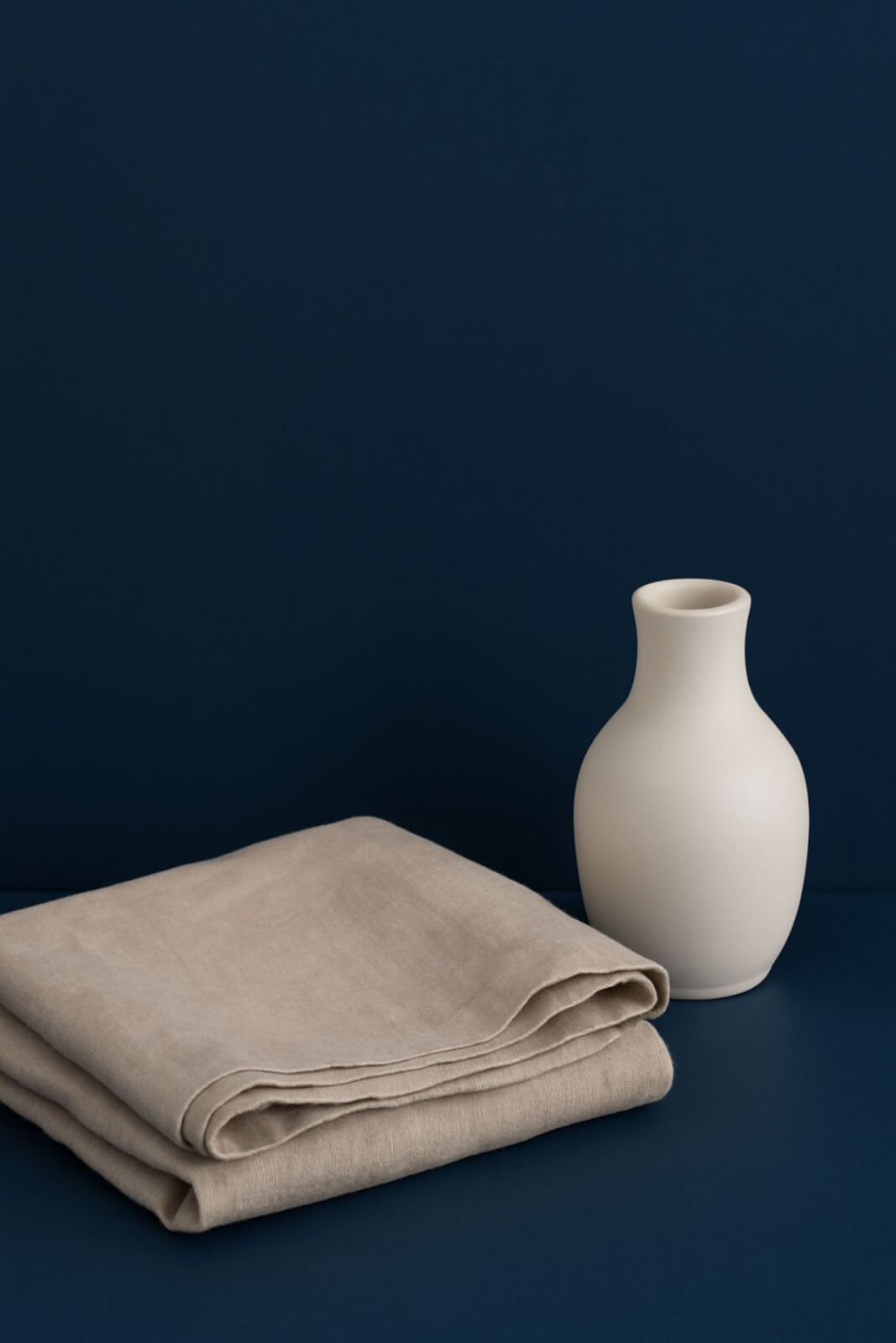 "Minimalist home decor scene with a folded beige linen throw and a smooth white ceramic vase against a deep blue background."